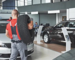 Monteur in rood shirt en grijze werkoverall draagt een autoband op zijn schouder in een moderne autoshowroom. Op de voorgrond staan een witte en een zwarte personenauto opgesteld, terwijl op de achtergrond medewerkers aan bureaus werken. De afbeelding straalt onderhoud, bandenservice en automotive dienstverlening uit.